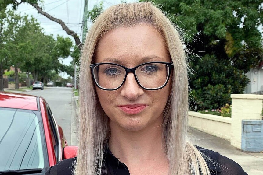 Megan Daniel, wearing black glasses, stands outside beside a car in the street.