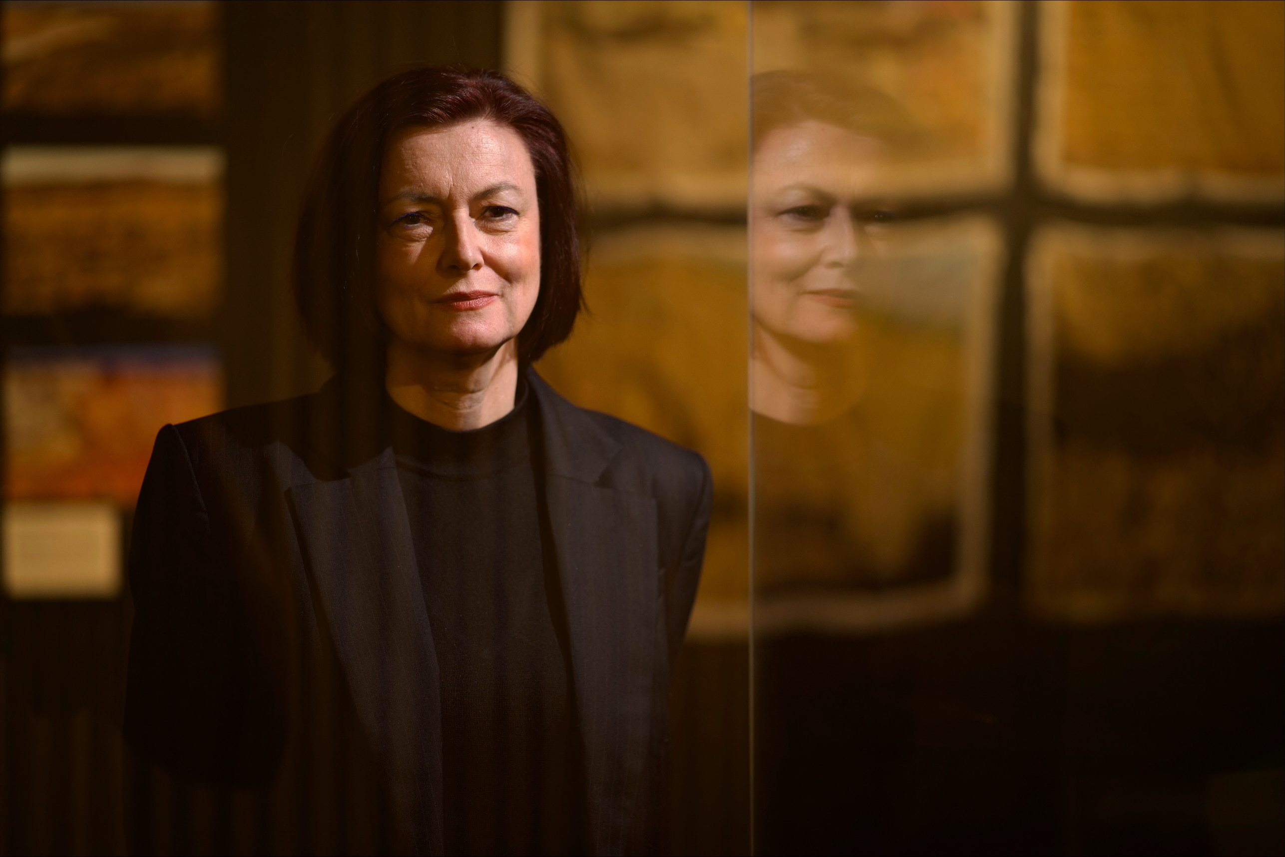 Kate Gould wears a black blazer and shirt. Artwork is hung behind her. She stands next to her reflection on the right.