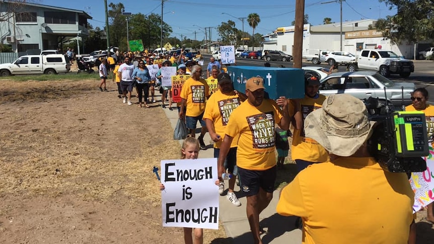 A crowd walks behind six men carrying a coffin painted blue. A child holds a sign reading ‘enough is enough’.