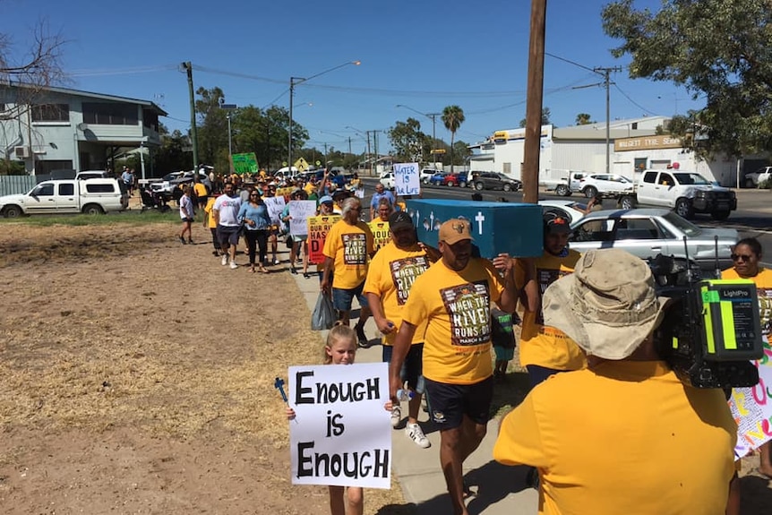 A crowd walks behind six men carrying a coffin painted blue. A child holds a sign reading ‘enough is enough’.