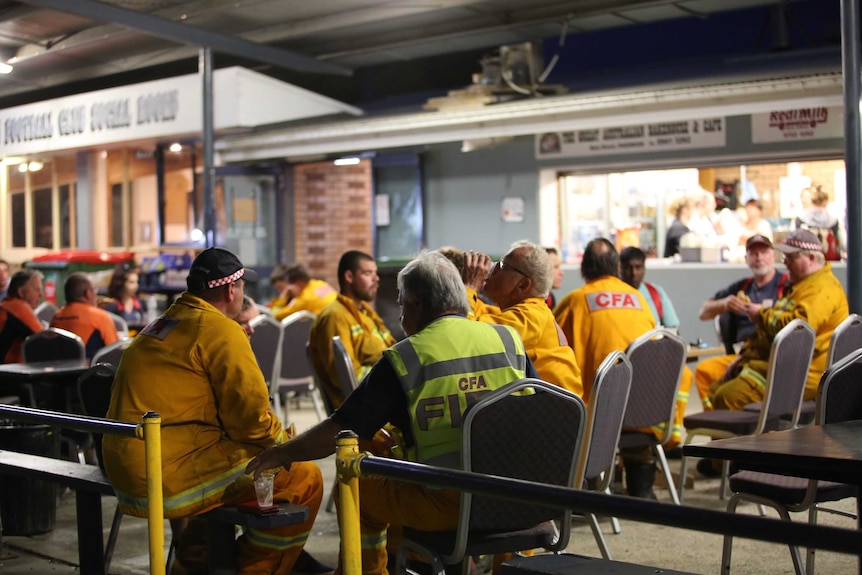 A group of firefighters wearing high visibility gear sit together while eating and drinking
