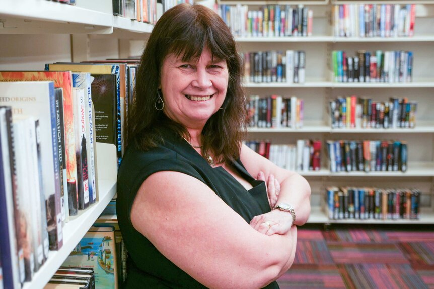 A middle-aged woman with brown hair and a fringe leans against library shelves, smiling.