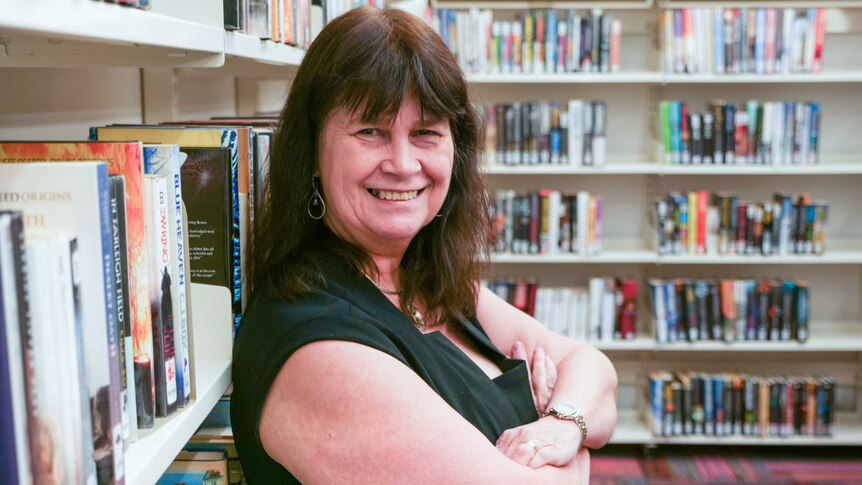 A middle-aged woman with brown hair and a fringe leans against library shelves, smiling.