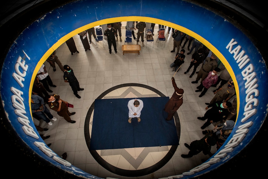 A view from above of one in the middle of a mat with one man standing whipping him.