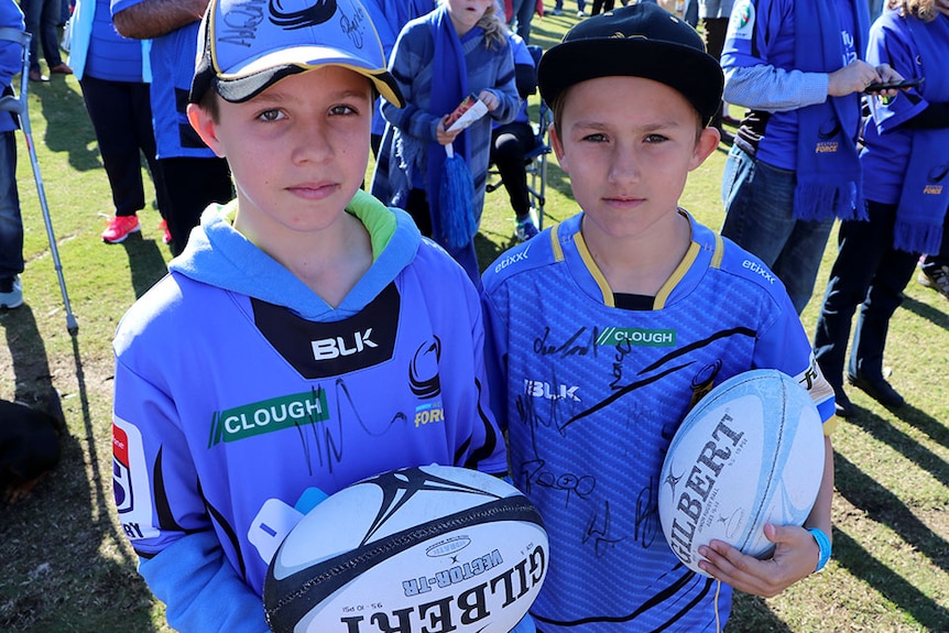 Two young boys dressed in Western Force uniform hold rugby balls, looking sad.