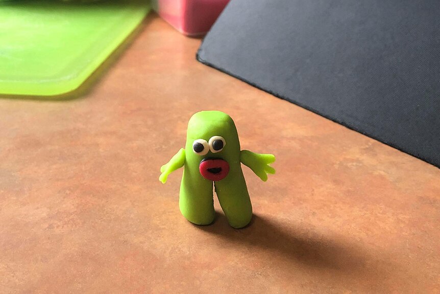 Little green man created out of modelling compound stands on a brown table.  