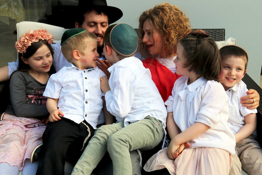 Rabbi Shmueli Feldman sitting on an outside chair with his wife, Rebbetzin Chasia Feldman, and their five children.