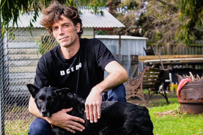 A man crouches down next to his black dog in a backyard. 