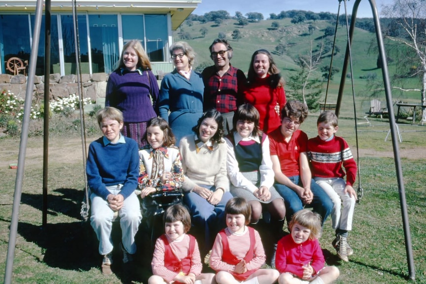A family photo of 11 children and mum and dad lined up in three rows at an outback property of green hills surrounding property