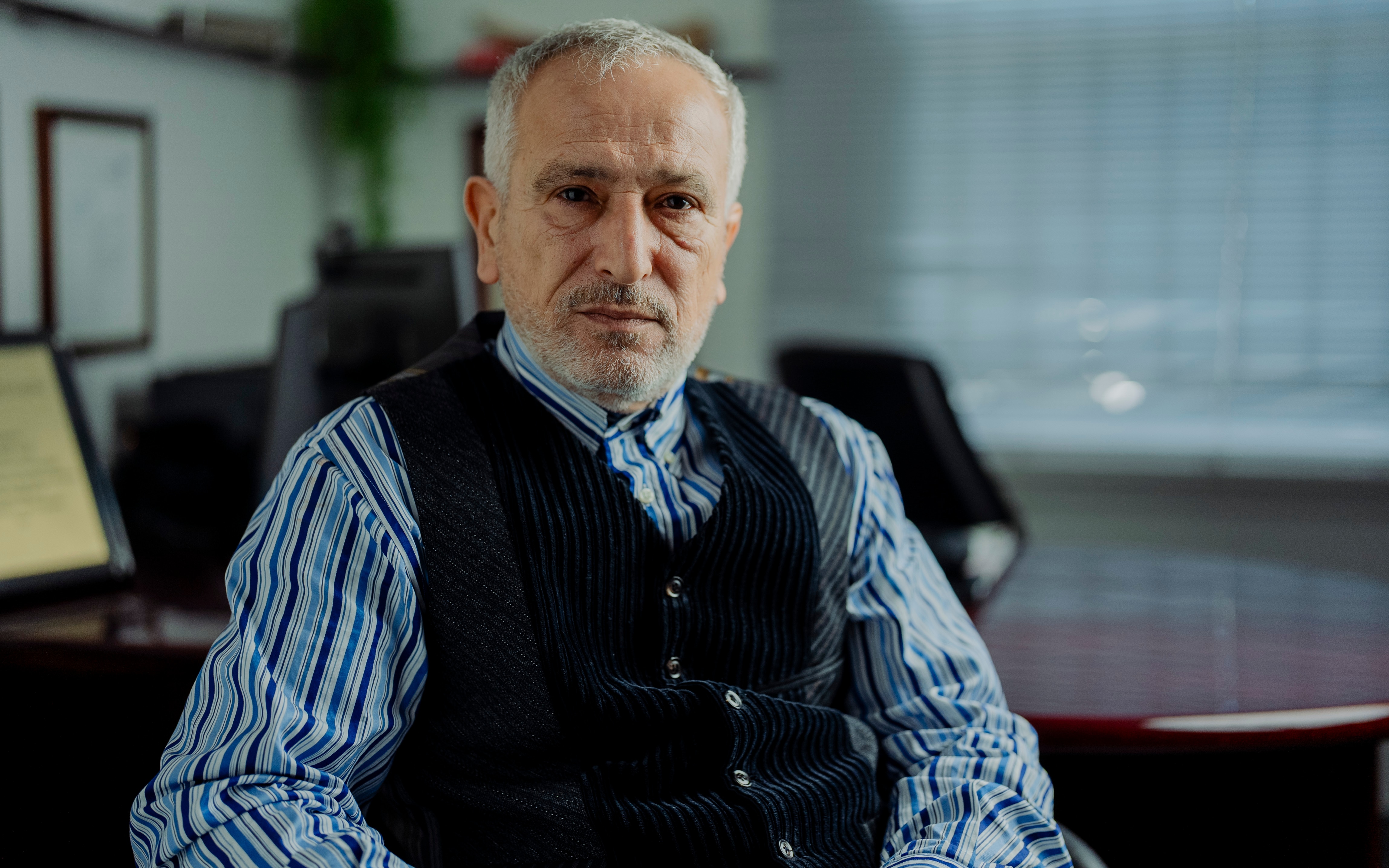 A man with grey hair sits in his office with a serious facial expression.