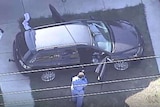 A person in a blue jumpsuit takes pictures of the inside of a black sedan parked on a lawn.