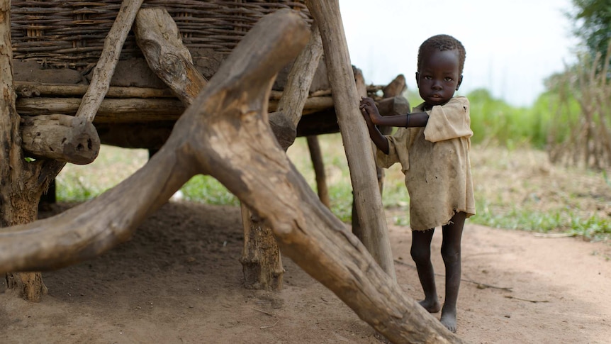 A thin child in South Sudan stands next to his family's grain store.