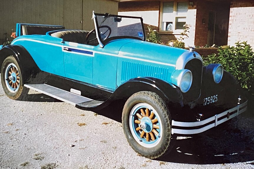 Aqua blue 1920s vintage car with wooden spoke wheels, black grill parked outside a house