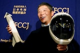 A man smiles as he holds a space helmet and model rocket up to be photographed.