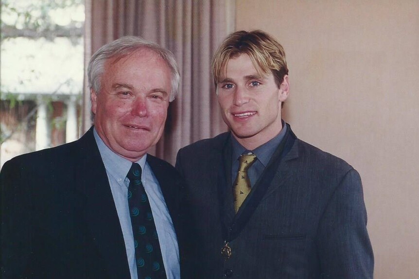 An older man in a suit next to a younger, blonde man in similar attire.