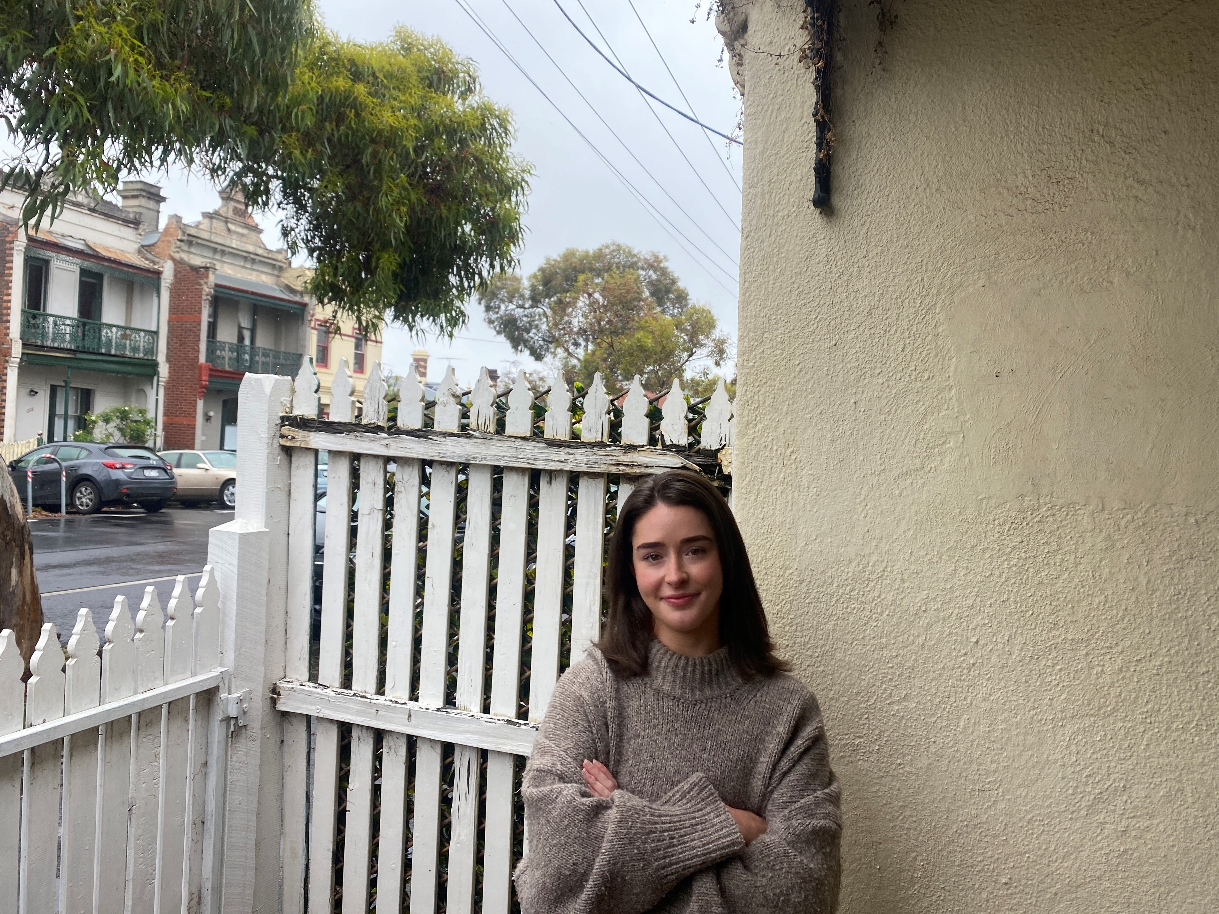 A woman wearing a light brown jumper stands next to a white picket fence.