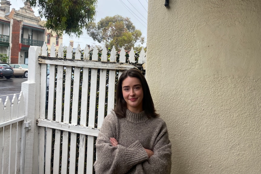 A woman wearing a light brown jumper stands next to a white picket fence.