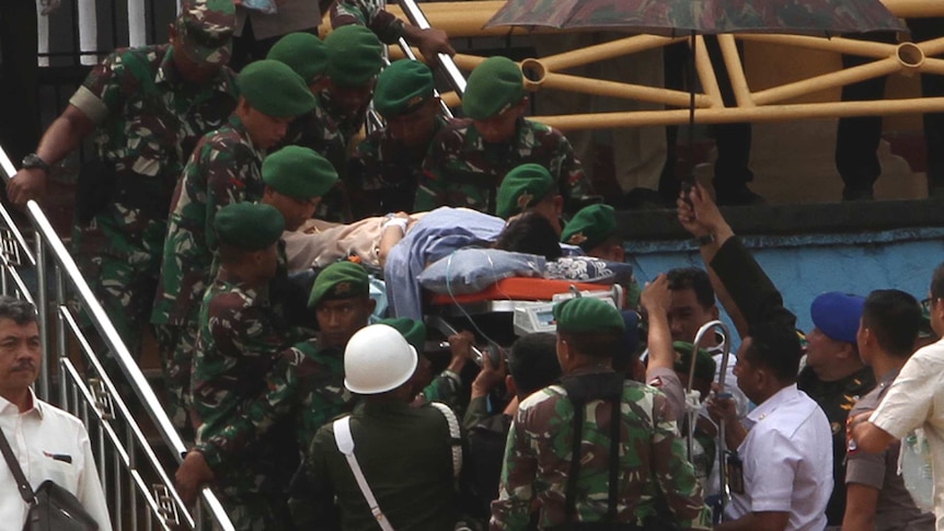 A number of soldiers wearing green camouflage carry a man on a stretcher up some stairs. One other man holds an umbrella.