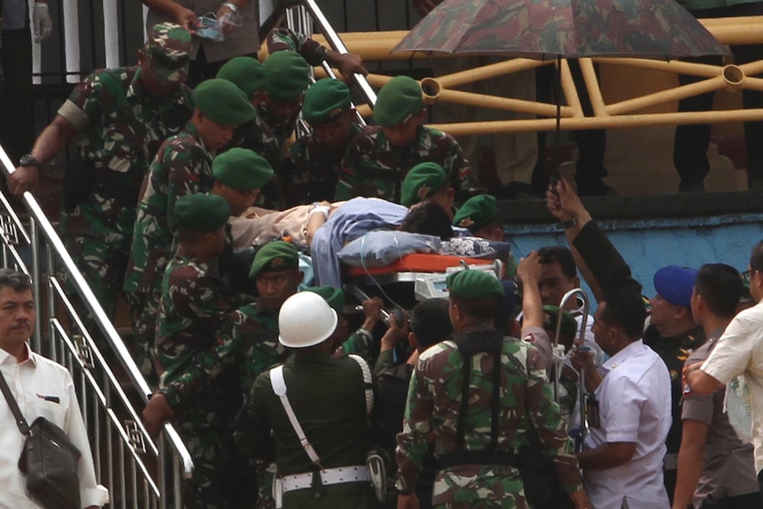 A number of soldiers wearing green camouflage carry a man on a stretcher up some stairs. One other man holds an umbrella.