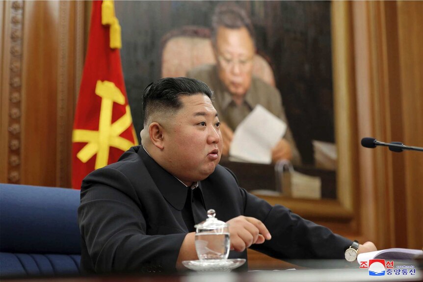 North Korean leader Kim Jong-un sits at a desk with a red and yellow flag in the background and an oil painting.