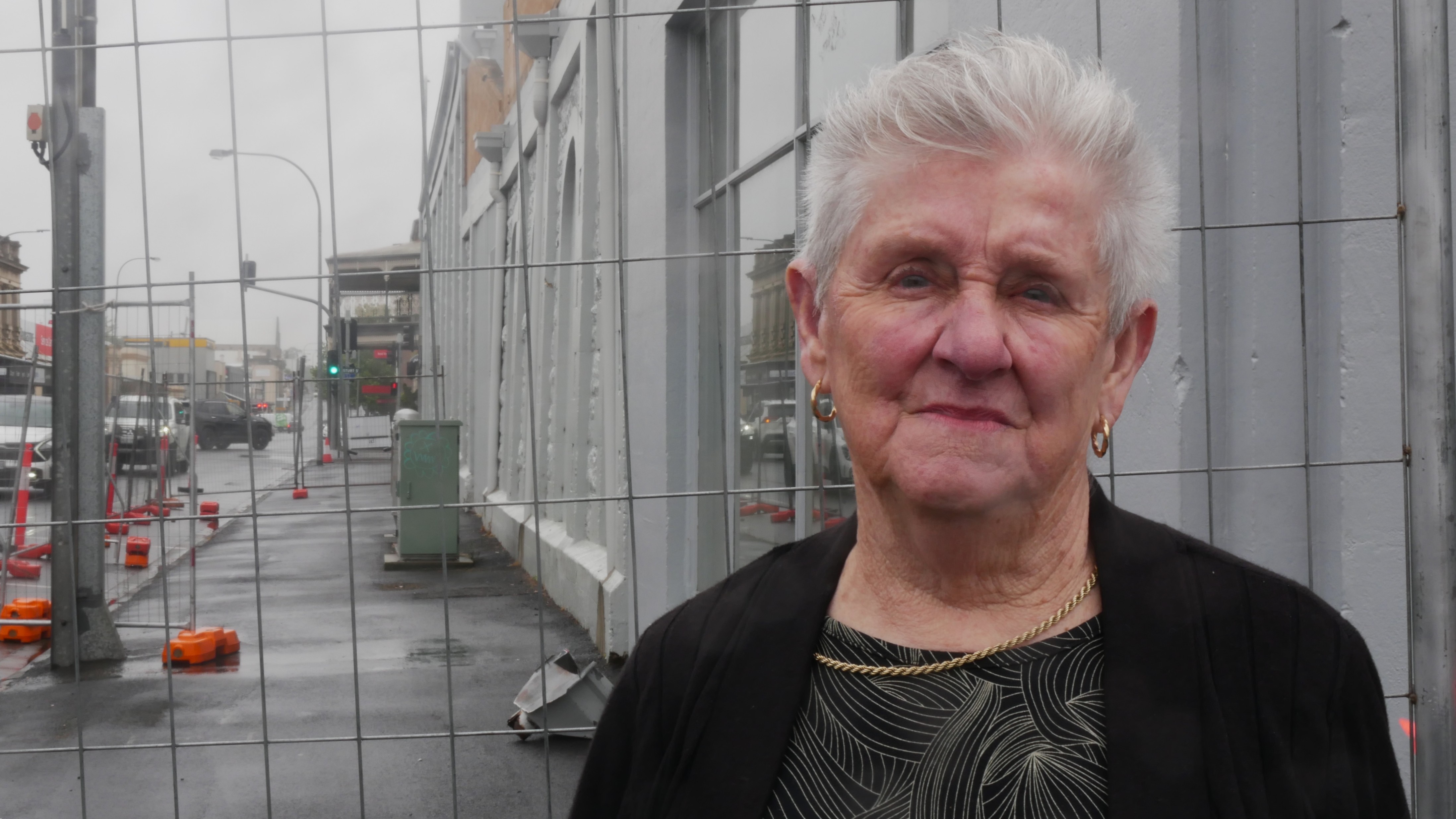 Uma mulher mais velha, de cabelos grisalhos, está em frente a uma cerca com uma parede azul-acinzentada com uma deformação significativa atrás dela.