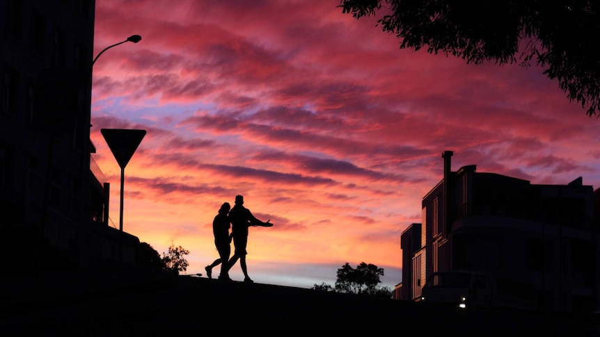 A pink and orange sunset in Australia, with two people and buildings silhouetted in front of it
