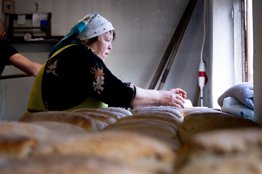 A woman in a headscarf kneads dough next to a row of freshly baked loaves of bread