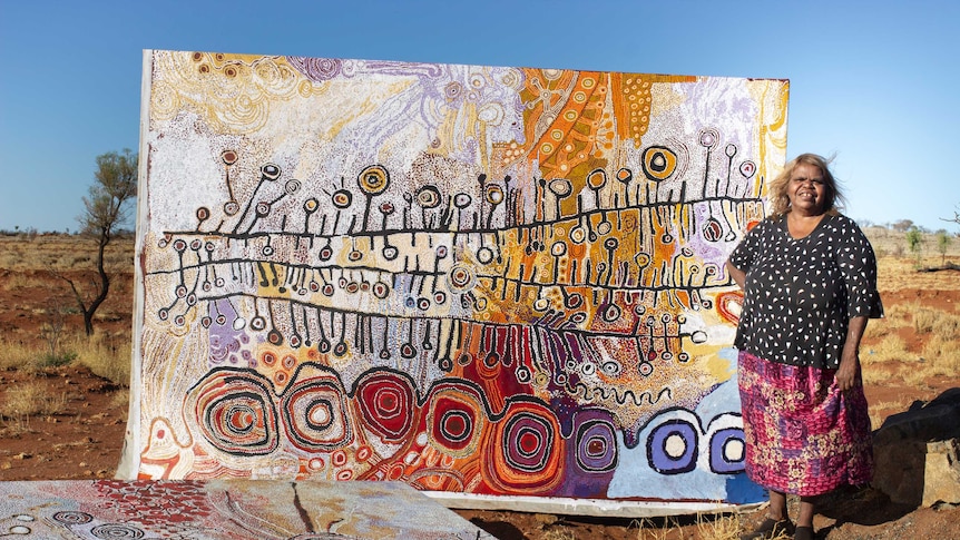 An Aboriginal woman stands in front of a large canvas in the Outback