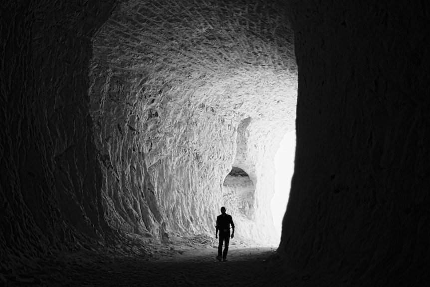 A black and white photo shows a male figure, in silhouette, walking through an underground tunnel. Light is seen at the far end.