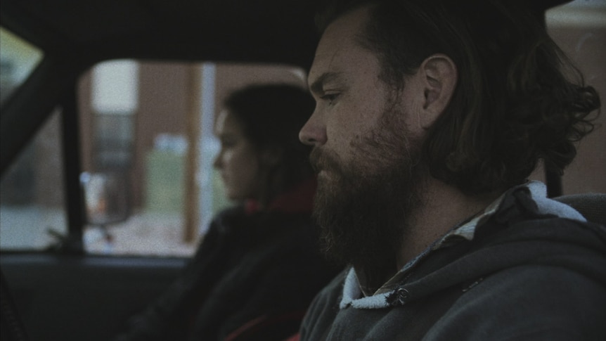 A man with beard and unruly hair sits in the front of a pick up truck, a woman sits next to him