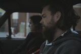 A man with beard and unruly hair sits in the front of a pick up truck, a woman sits next to him