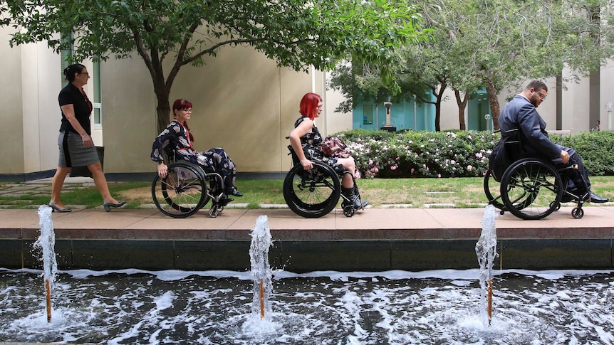 A man and two women push their wheelchairs through a courtyard alongside water fountains. A woman walks behind them