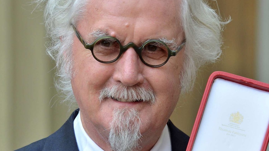 Billy Connolly wears glasses and a suit and holds up a medal