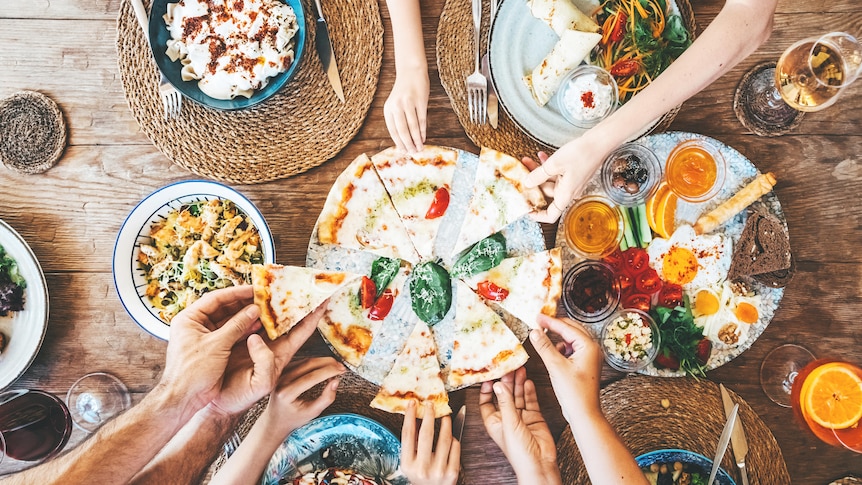 Picture of wooden table with colourful food dishes, pizza in the centre and hands reaching out to grab a slice