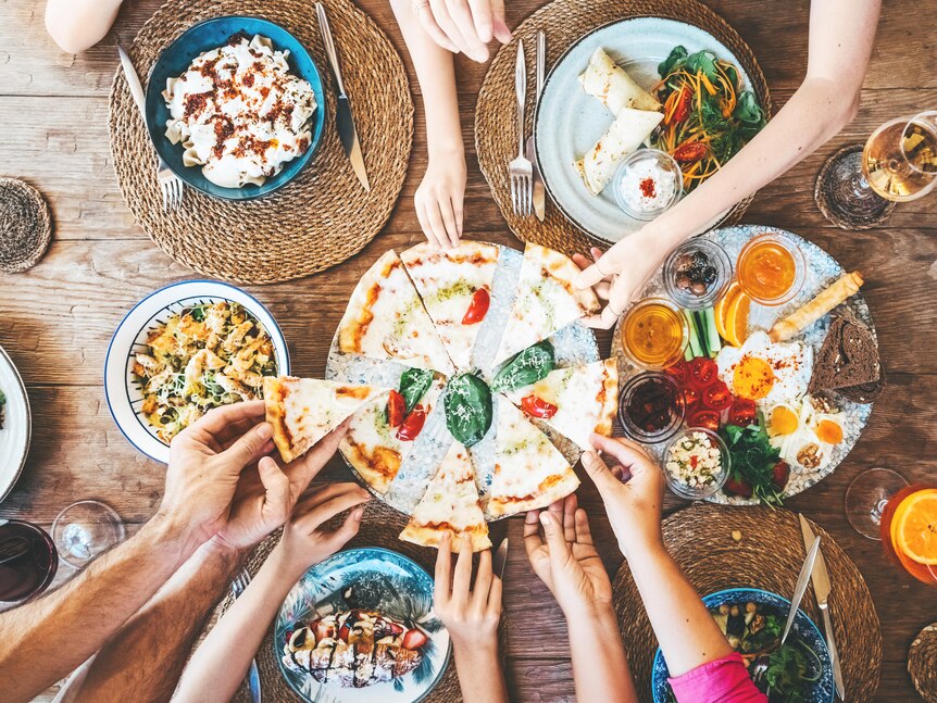 Picture of wooden table with colourful food dishes, pizza in the centre and hands reaching out to grab a slice