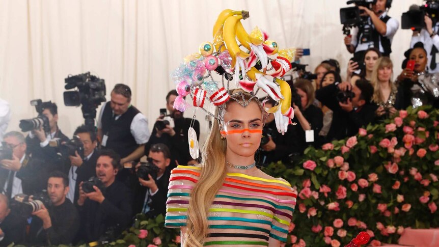 Cara Delevingne wears bananas on her head with a translucent and rainbow striped outfit, complete with a cane.