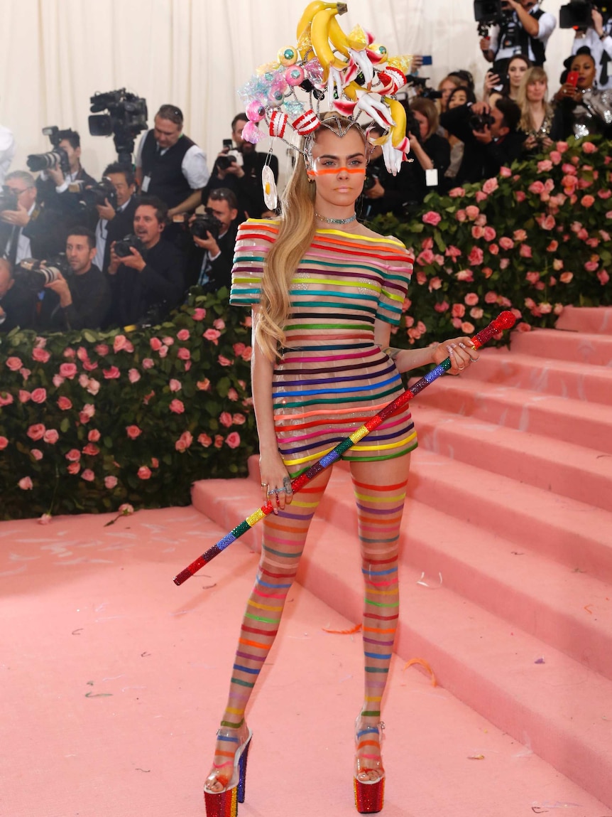 Cara Delevingne wears bananas on her head with a translucent and rainbow striped outfit, complete with a cane.