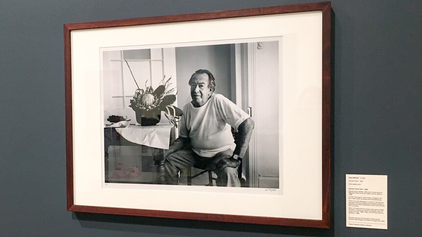 A black and white photograph of Donald Friend hanging on the wall of an art gallery.
