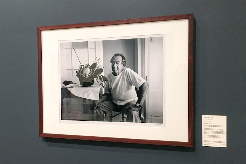 A black and white photograph of Donald Friend hanging on the wall of an art gallery.