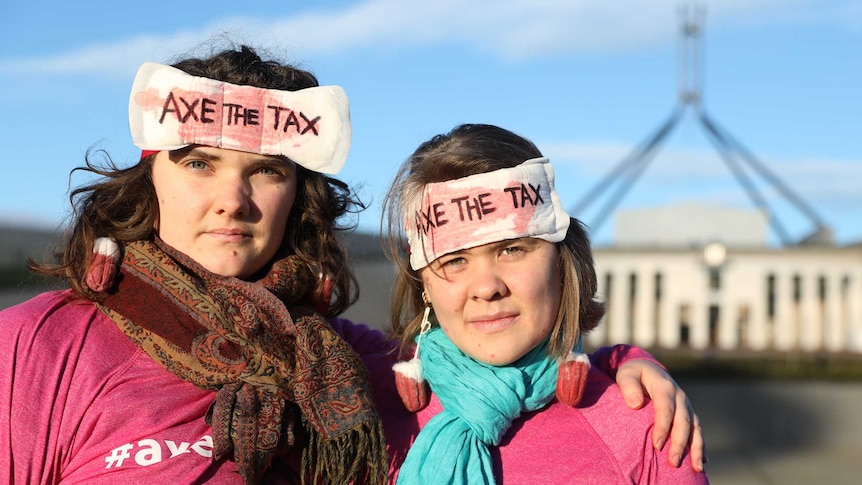 Two women pose with serious expressions, wearing tampon earrings and sanitary pads that say 'AXE THE TAX' stuck to their heads.