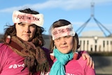 Two women pose with serious expressions, wearing tampon earrings and sanitary pads that say 'AXE THE TAX' stuck to their heads.