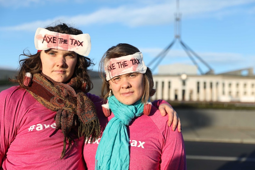 Two women pose with serious expressions, wearing tampon earrings and sanitary pads that say 'AXE THE TAX' stuck to their heads.