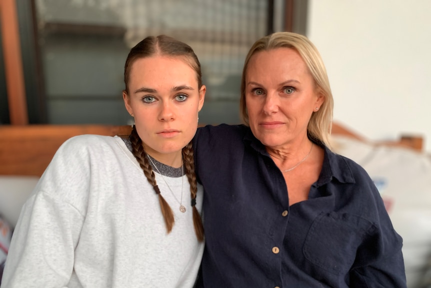 A young woman sits beside her mother on a couch. Both look into camera with serious expressions.