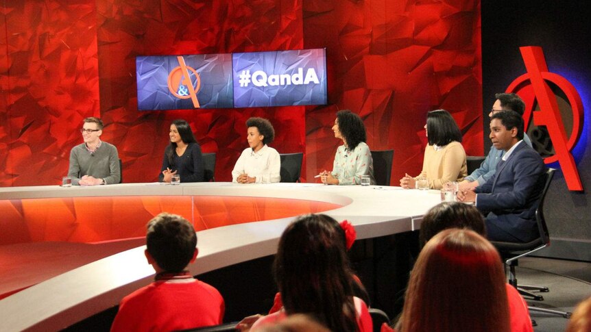 Seven people sitting behind the Q&A desk in a television studio.