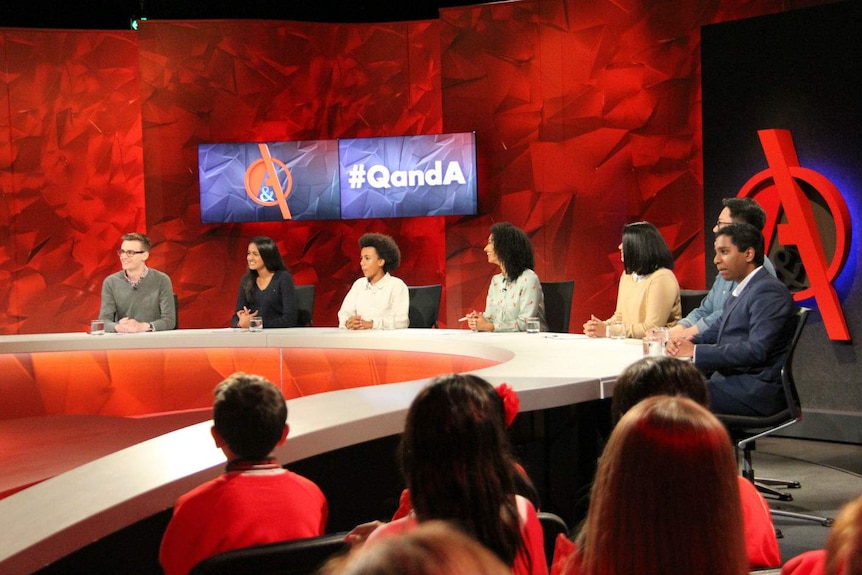 Seven people sitting behind the Q&A desk in a television studio.