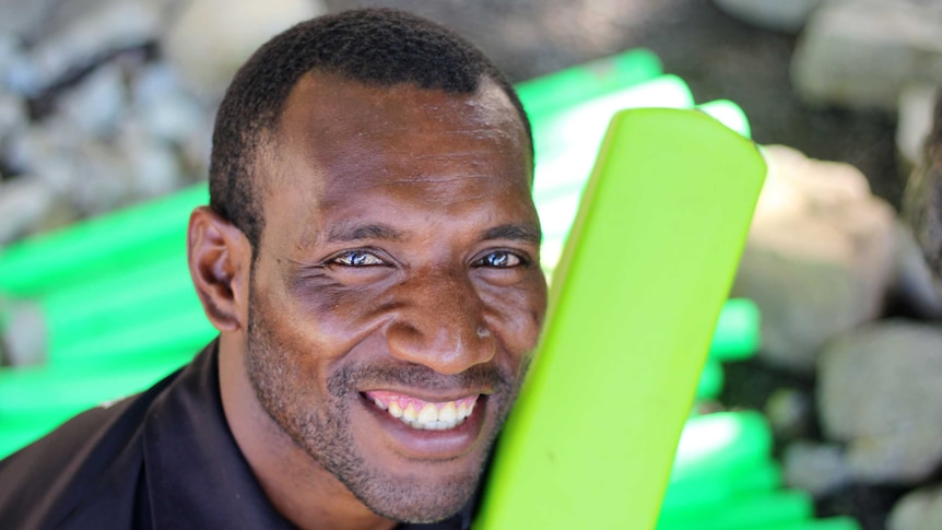 A PNG man with short-cropped hair smiles a camera holding a green plastic cricket bat over his shoulder.