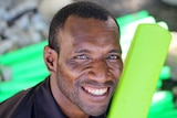 A PNG man with short-cropped hair smiles a camera holding a green plastic cricket bat over his shoulder.
