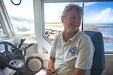 Female skipper sits behind the wheel of her boat with the blue sky and ocean visible through the window behind her