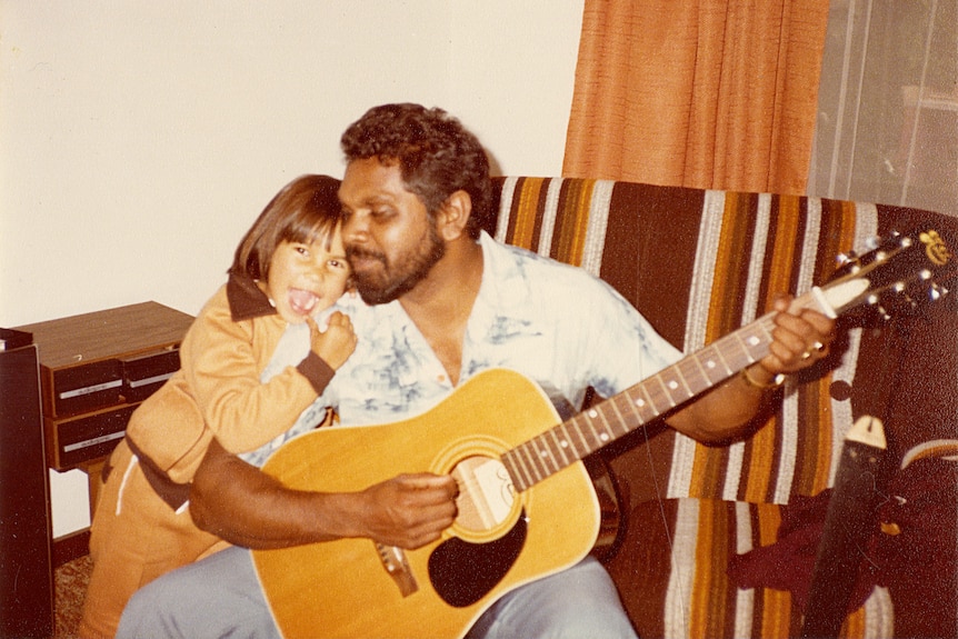 A sepia toned photo shows a young girl hugging her bearded father, who is playing an acoustic guitar