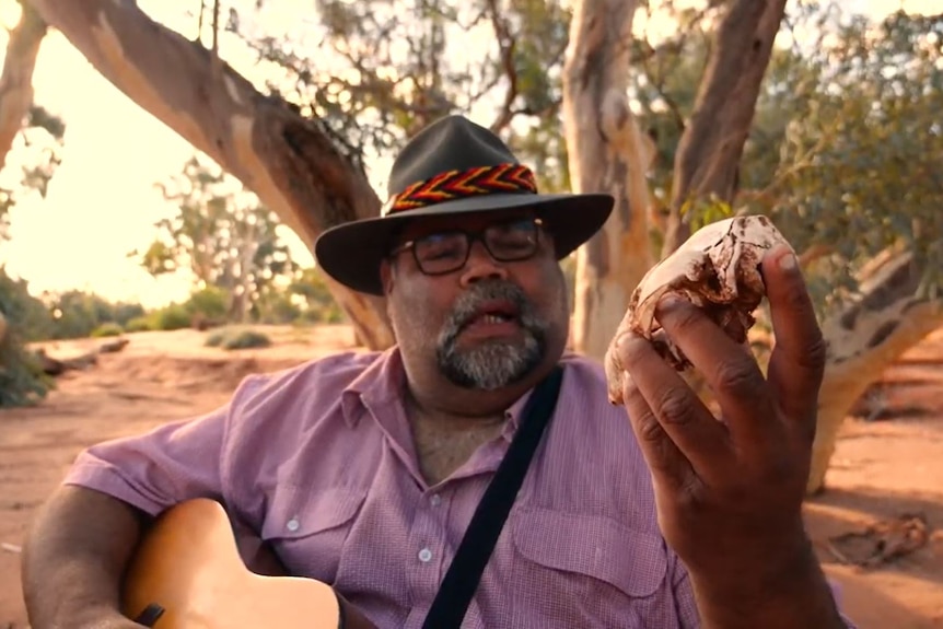 Un homme dans le désert avec un arbre dans le dos, tenant un totem avec une guitare autour de lui